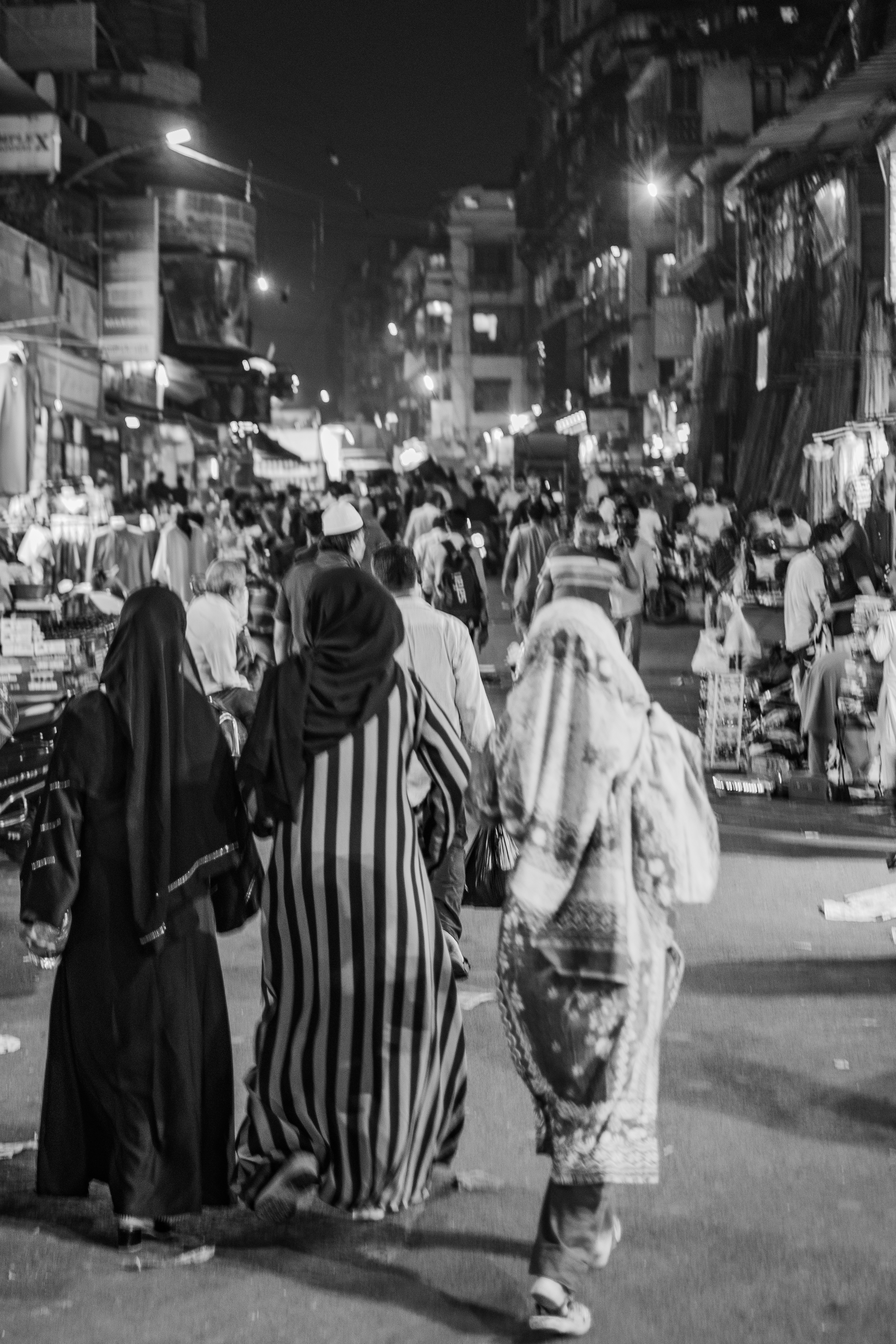 Three figures walking away from camera into the crowded street at night, B&W