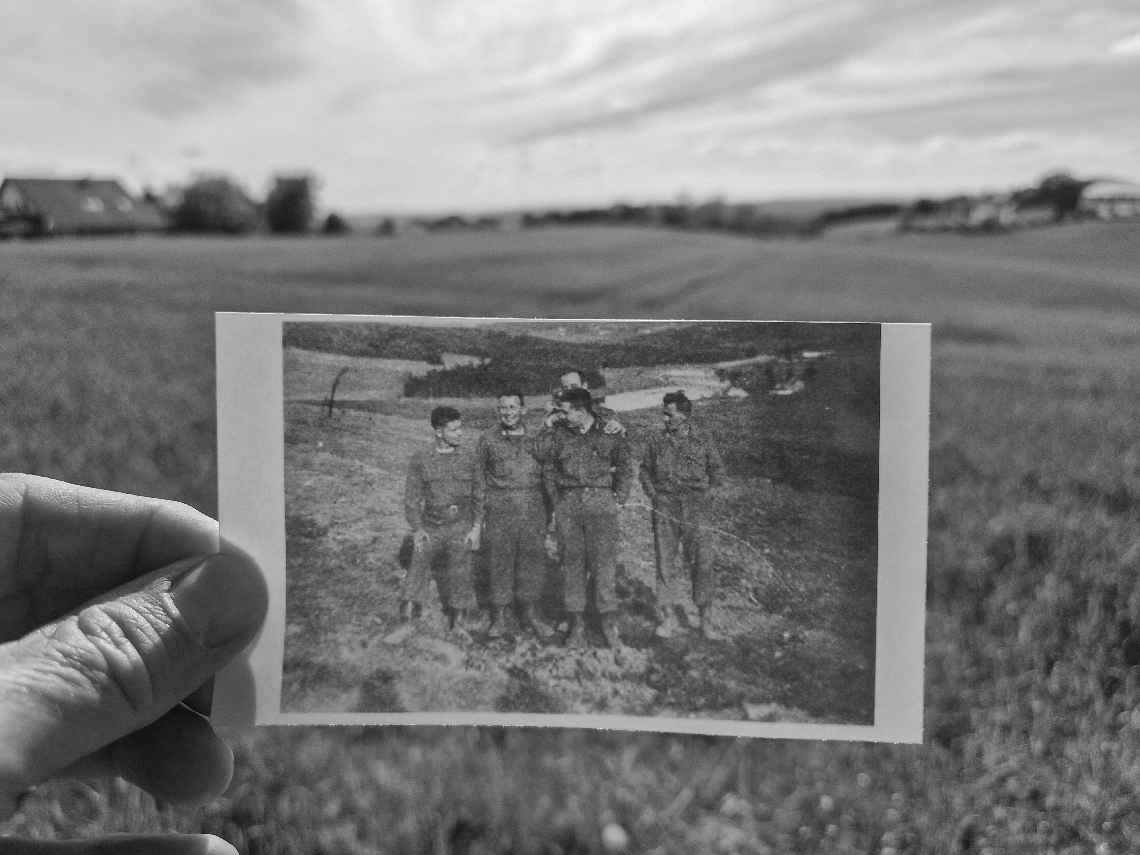 Hand holding a sepia VE Day photograph in front of the same field 81 years later