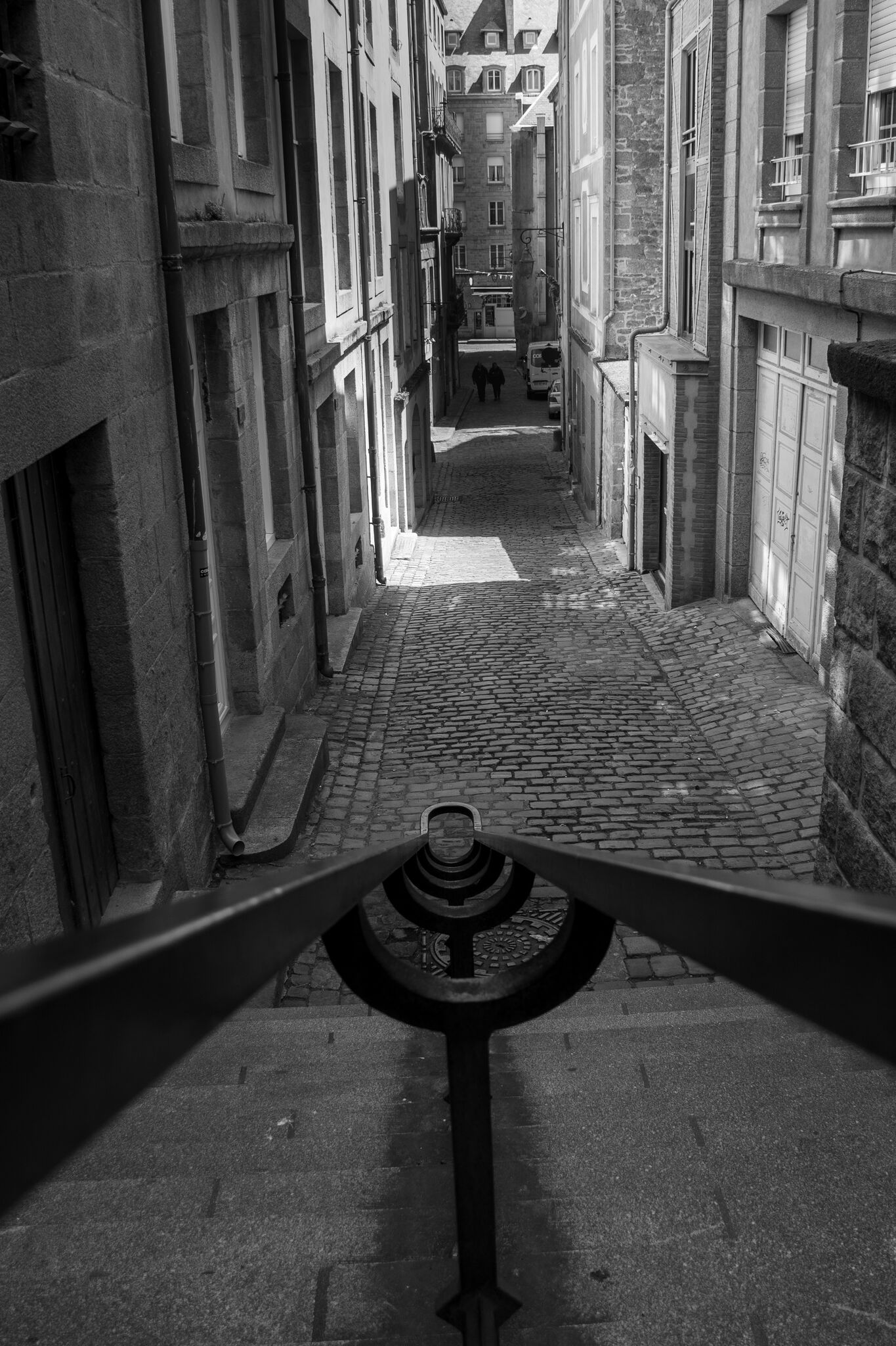 Looking down a staircase into a narrow cobblestone lane, two figures in the distance, black and white
