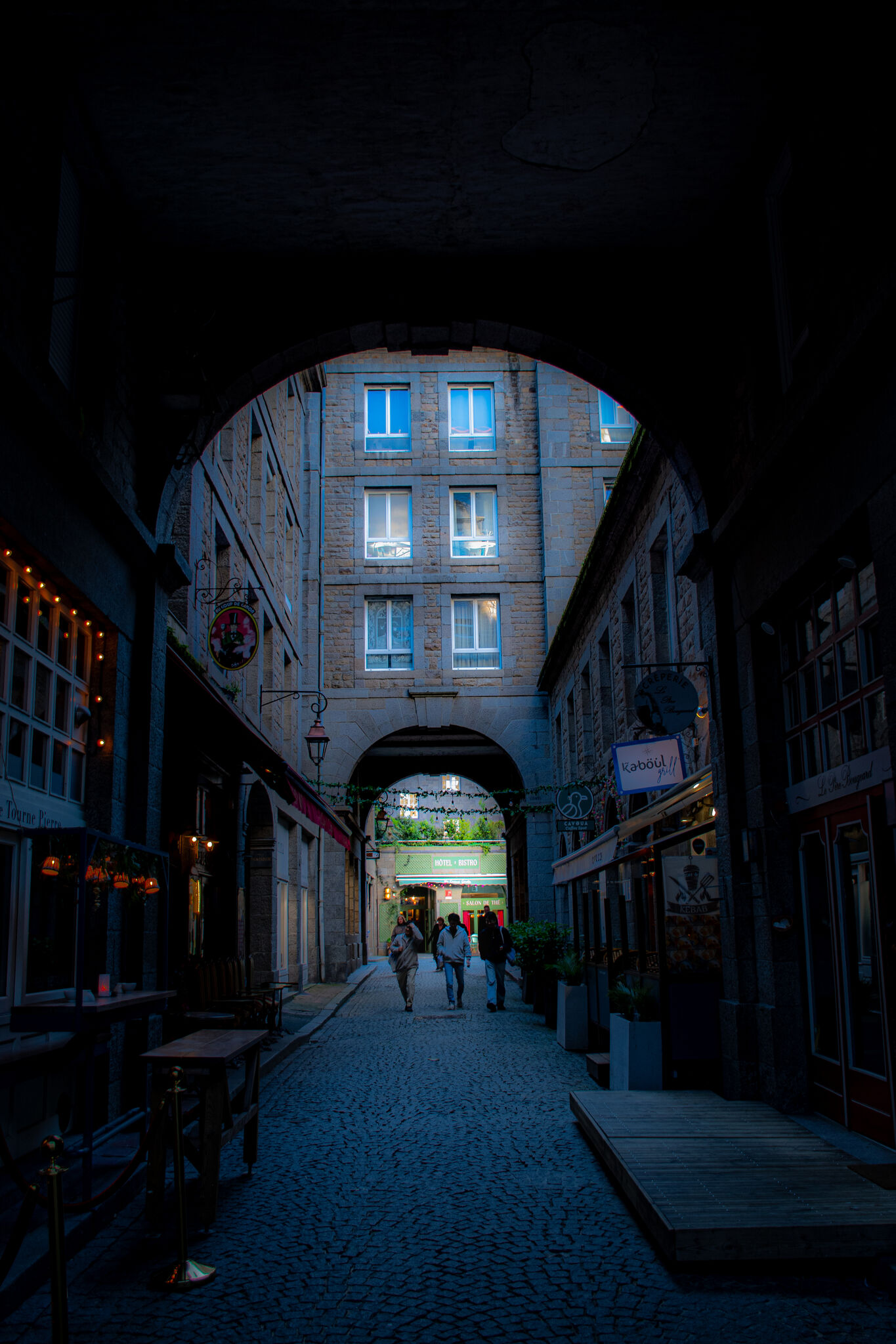 Looking through a stone archway into a lit street beyond, people walking at dusk