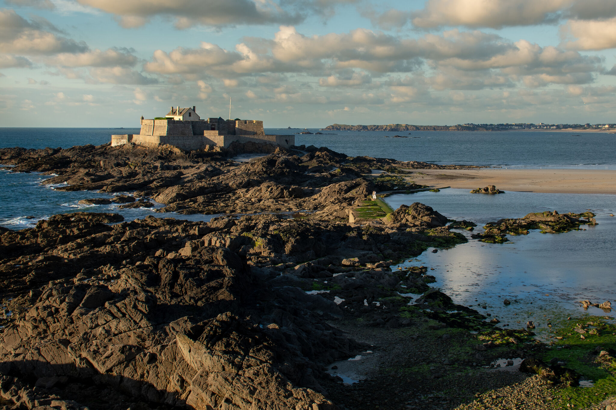 Fort National at golden hour, tidal flats glowing, single figure on the causeway